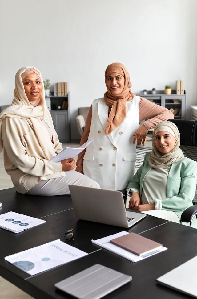 Three people wearing hijabs standing and sitting in an office, laughing. Image by Cedric Fauntleroy on Pexels.